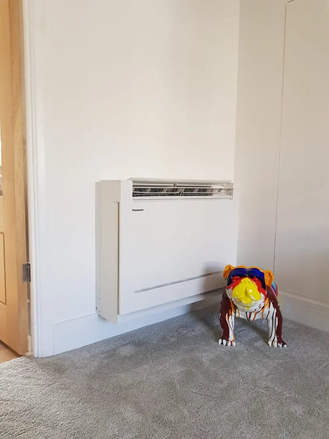 Floor-mounted air conditioning unit installed under window with colorful decorative item nearby in a home setting.